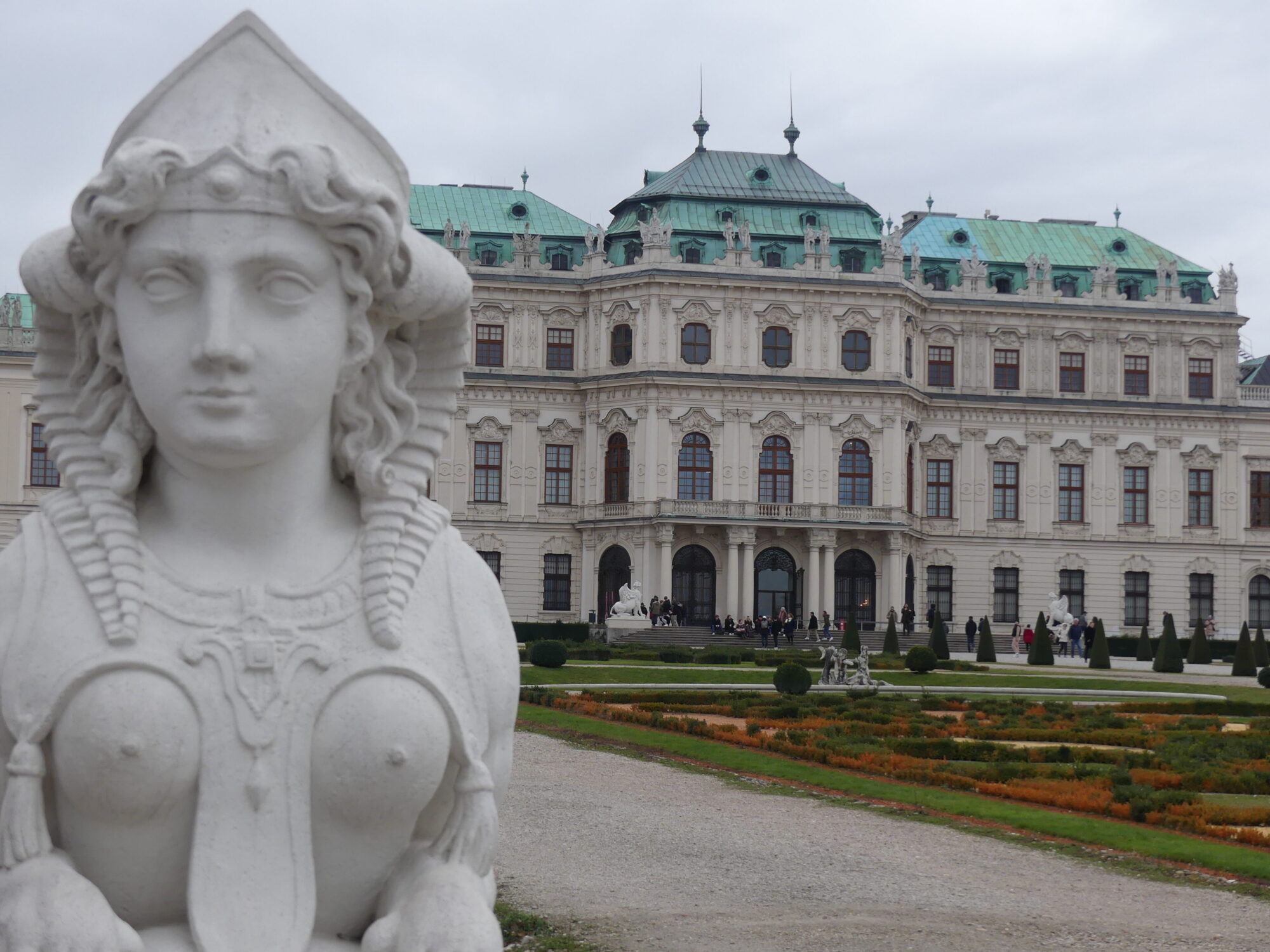 In the foreground a statue of a mermaid. In the second plan gardens. In the third plan, the palace with white façade and green roofs.