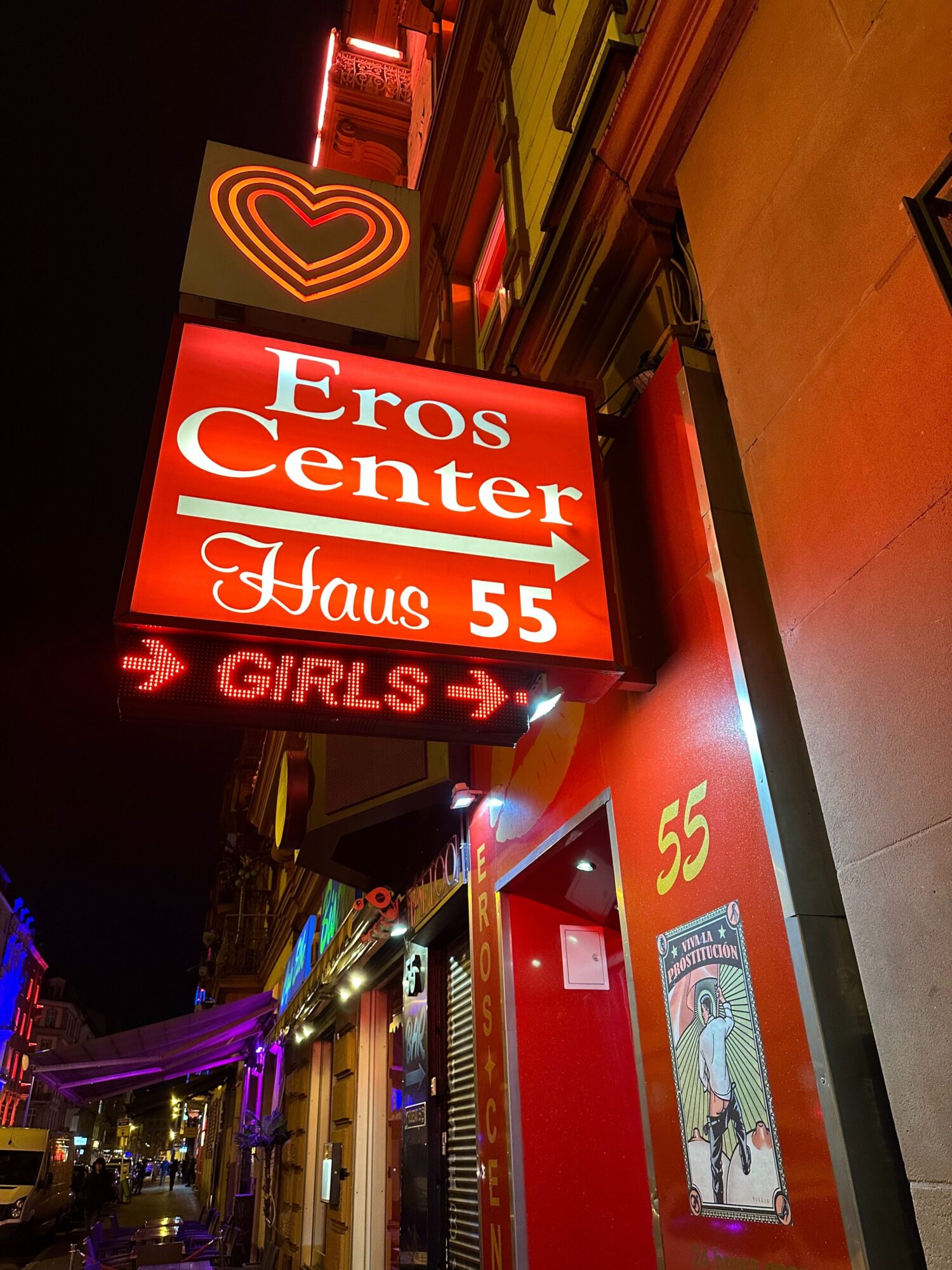 Gros plan sur un panneau indiquant "Eros Center Haus 55" et "Girls". 
Ce panneau est situé sur un petit immeuble rouge lui même situé dans une petite rue illuminée de nuit.