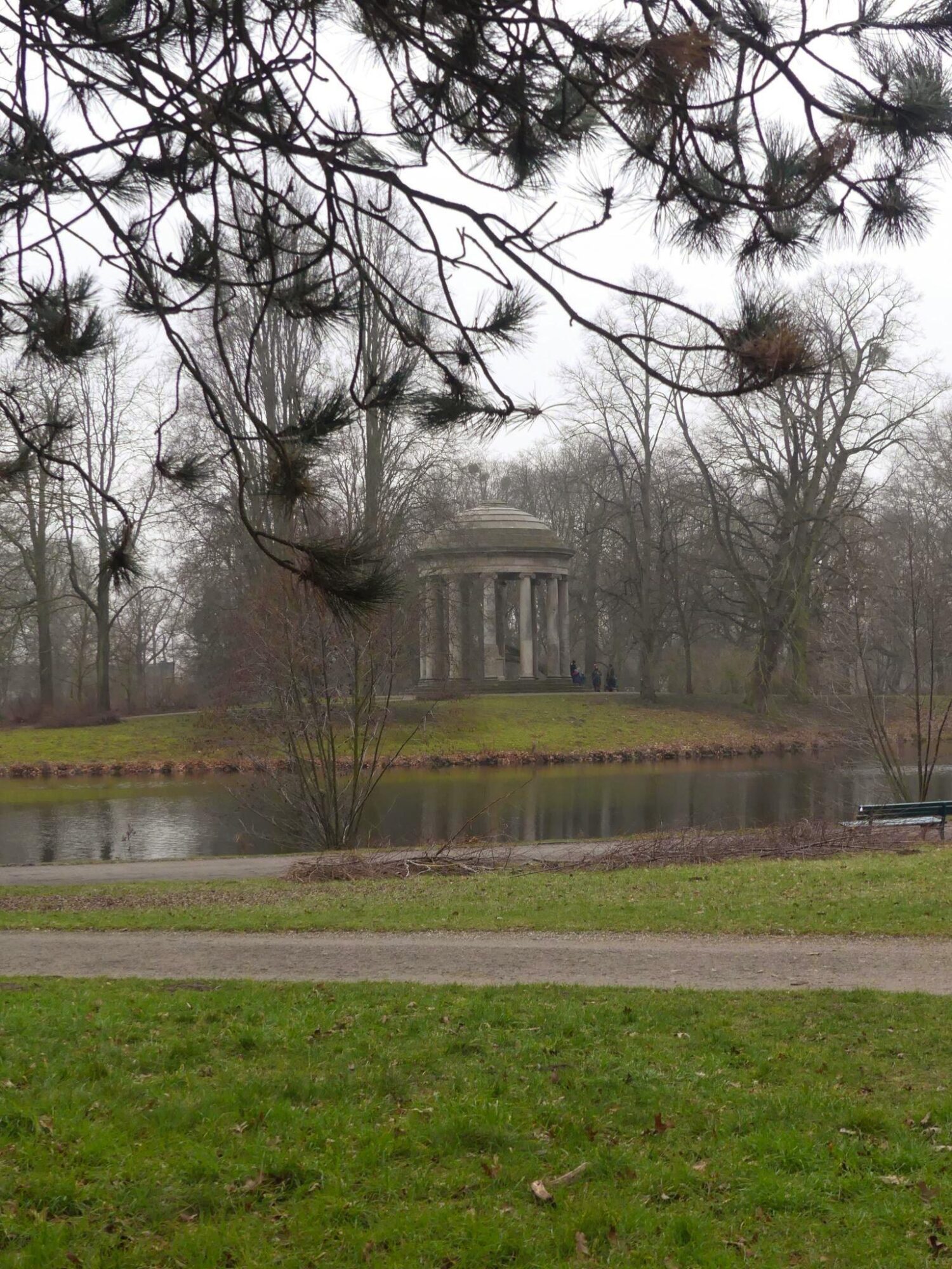 View of a park made up of trees without foliage and a small island of land in the middle of a pond. On the islet is a temple.